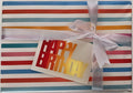 A wrapped gift box with colorful vertical stripes in red, blue, orange, and pink on a white background, tied with a white satin ribbon and a small gold foil 'Happy Birthday' card.